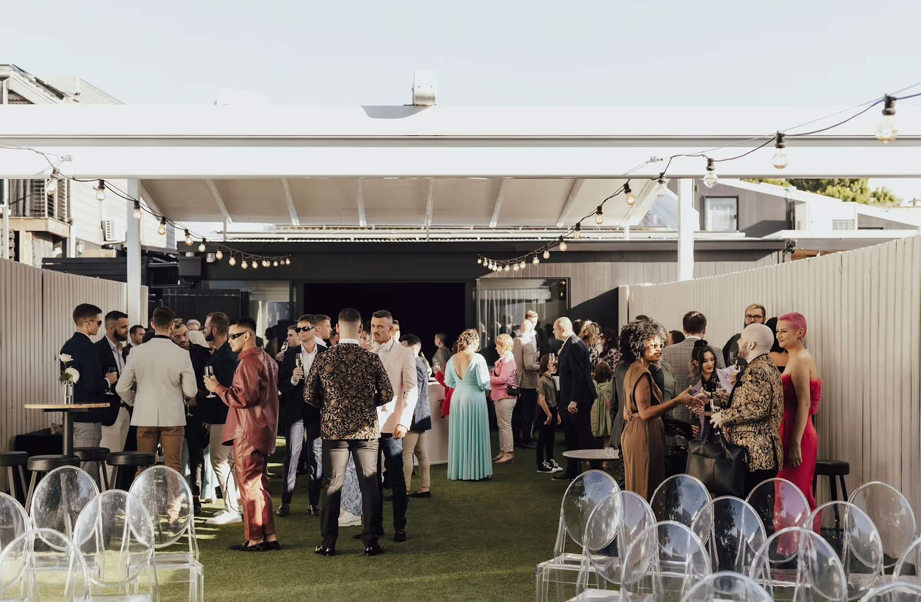 Wedding guests mingling in the private garden courtyard at The Refinery Brisbane with festoon lights and clear ceremony chairs.