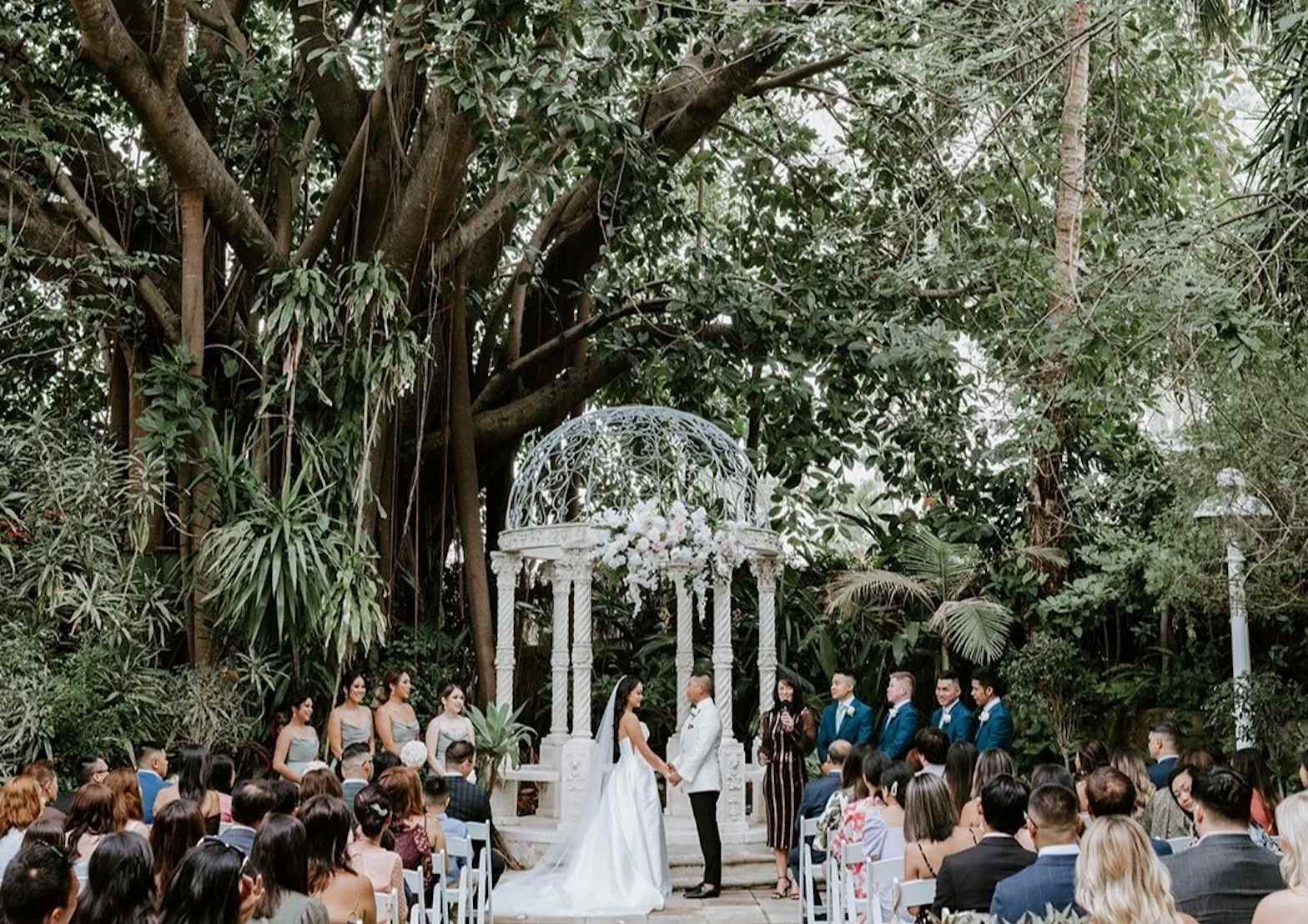 Garden wedding ceremony at Boulevard Gardens Brisbane with guests seated under tropical greenery and a white floral gazebo.