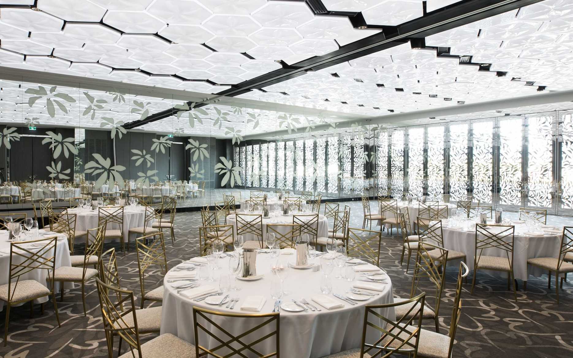 Wedding reception setup in the Frangipani Ballroom at Emporium Hotel South Bank with round tables, gold chairs and modern floral ceiling details.