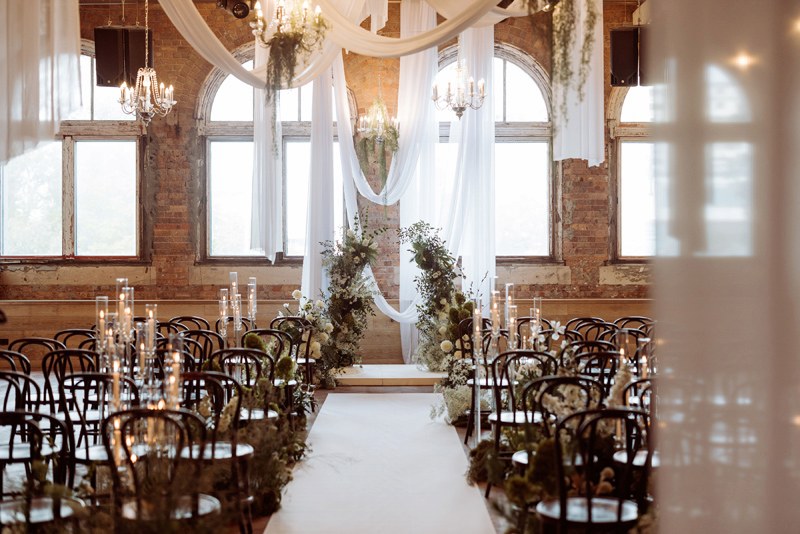 Wedding ceremony setup at The Warehouse Brisbane with exposed brick walls, chandeliers, draped fabric and floral aisle styling.
