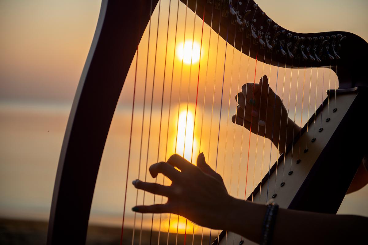 A close up of a lady hands playing a hard on the beach with the sunset in the background.