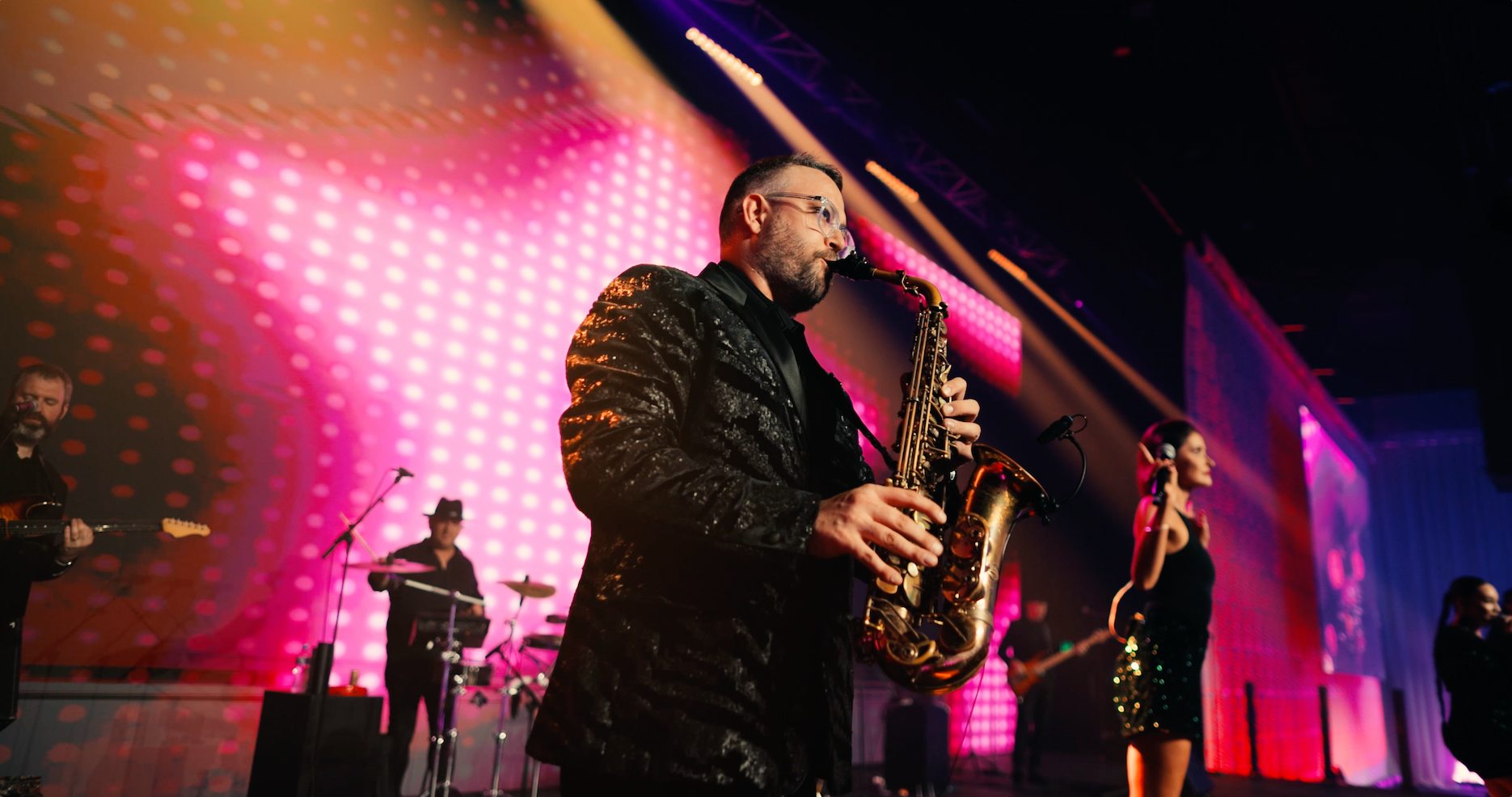 Corporate party saxophonist performing with a live band on stage under pink event lighting.