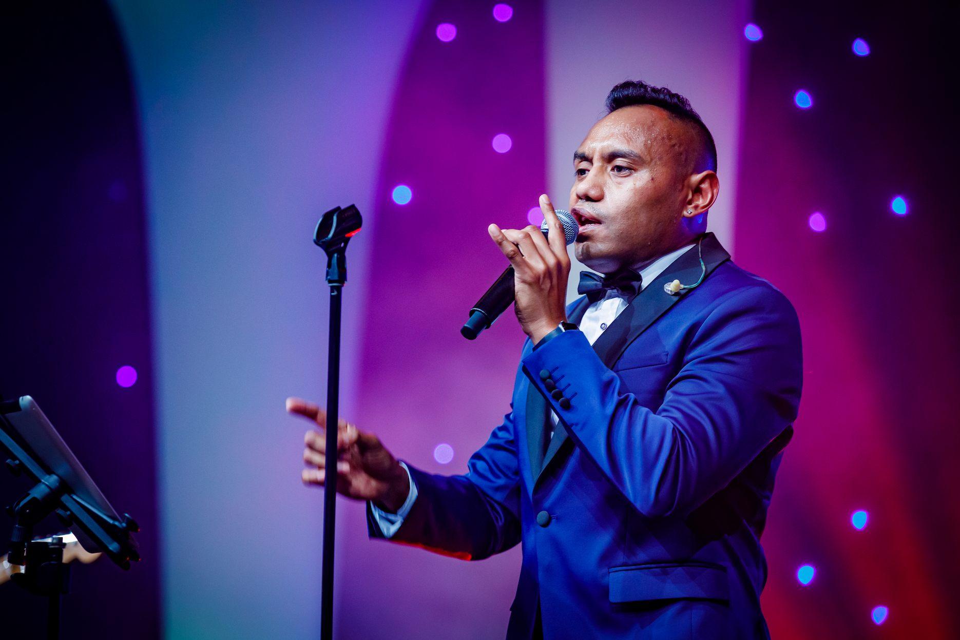 Live wedding singer in a blue tuxedo performing under vibrant purple and blue stage lighting.