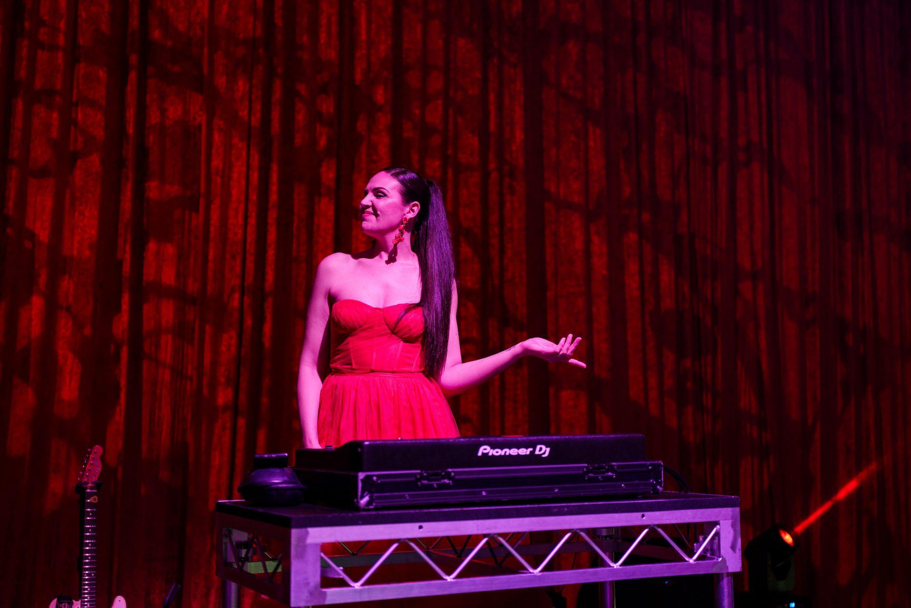 Female DJ performing behind Pioneer DJ decks under red stage lighting.