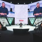 Australian keynote speaker presenting on stage at a corporate conference with large event screens and audience seating.