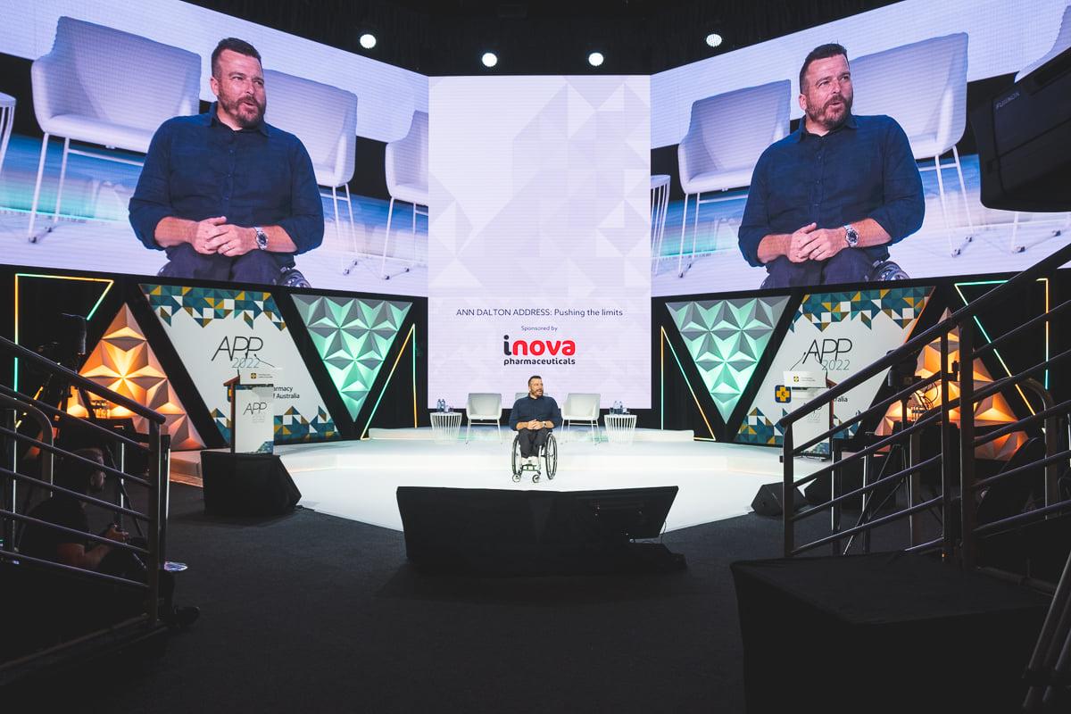 Australian keynote speaker presenting on stage at a corporate conference with large event screens and audience seating.