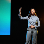 Female guest speaker presenting on stage at a corporate event with a headset microphone.