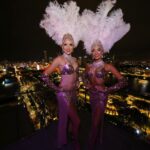Roving showgirls in sequinned costumes and feather headdresses at a rooftop corporate event.