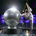 Acrobat performing beside a giant disco ball at a disco theme party.
