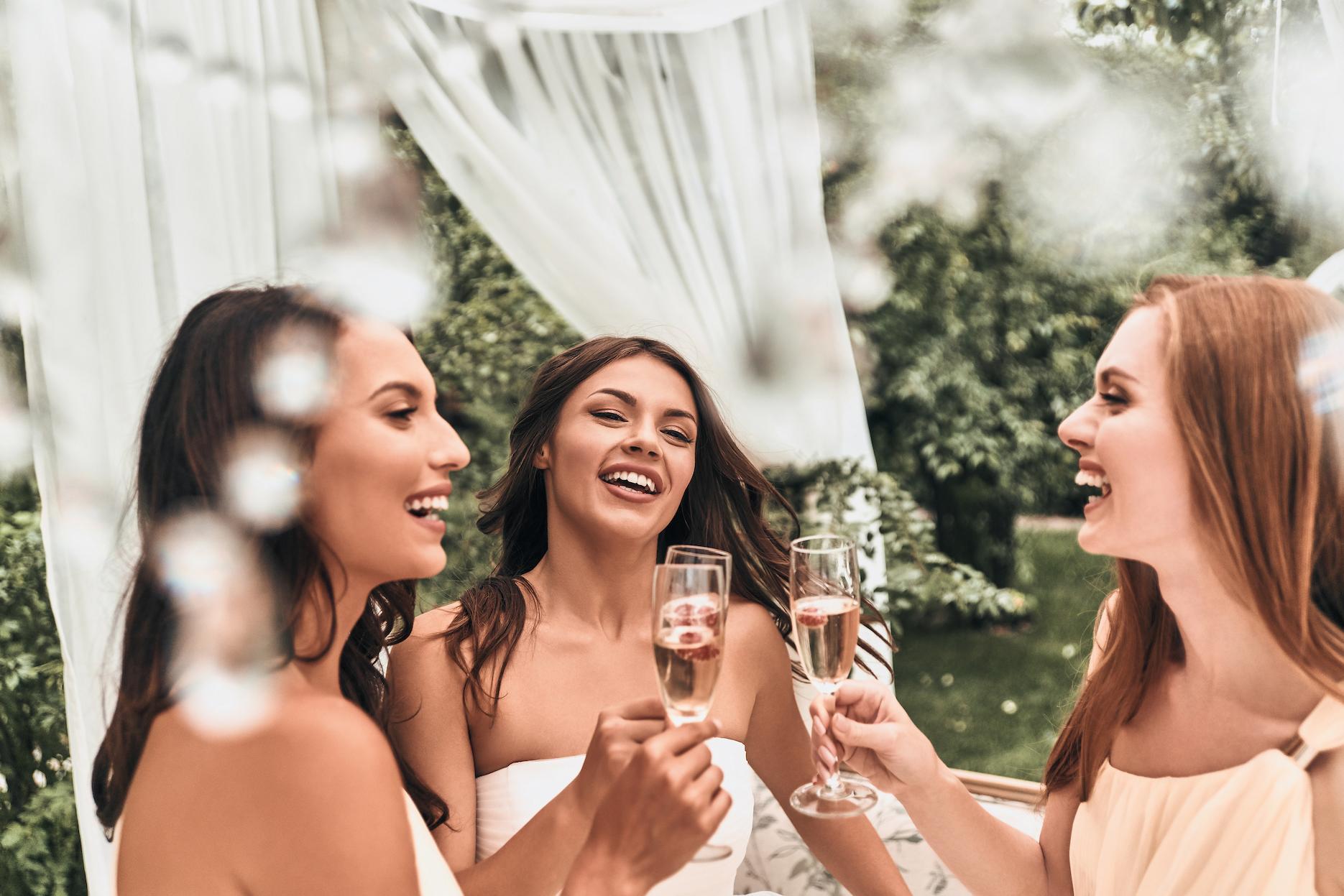 Three friends laughing and toasting champagne at an outdoor bridal shower celebration.