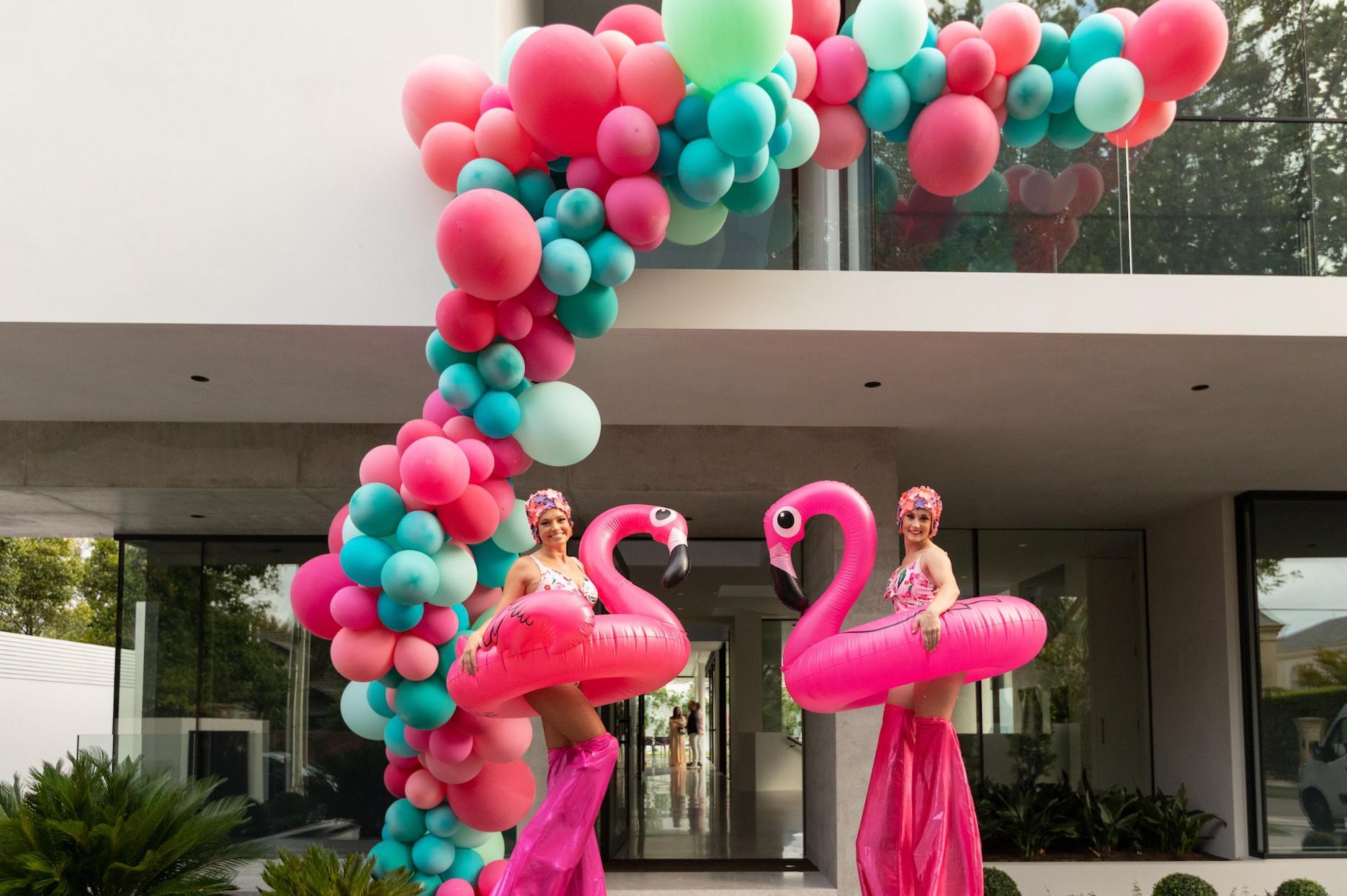 Roving flamingo performers under a balloon arch at a themed birthday party.
