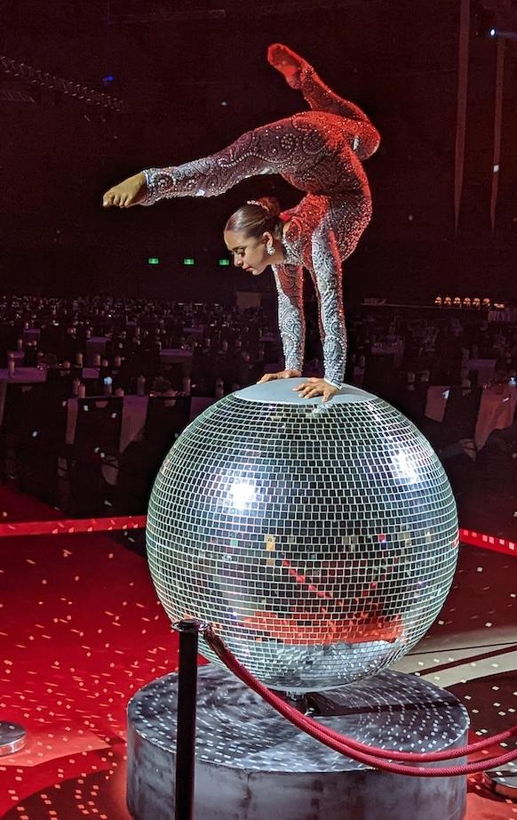 Contortionist performing a hand-balancing act on a giant mirror ball at a formal event.