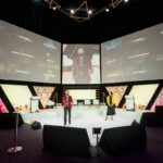 Corporate conference stage with presenters and large event screens.