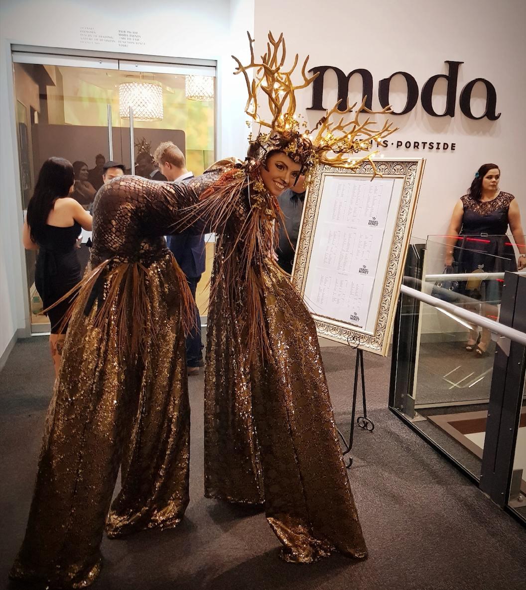 Onstage stilt walkers in elaborate gold costumes and antler headpieces welcoming guests at a corporate event.