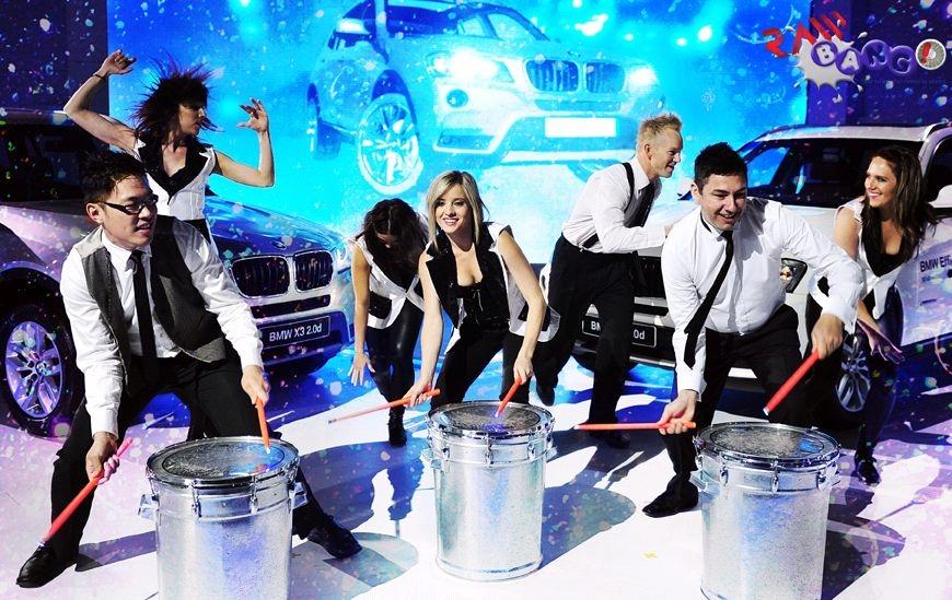 Onstage percussion performers and dancers delivering a high-energy branded feature act in front of BMW vehicles at a corporate event.
