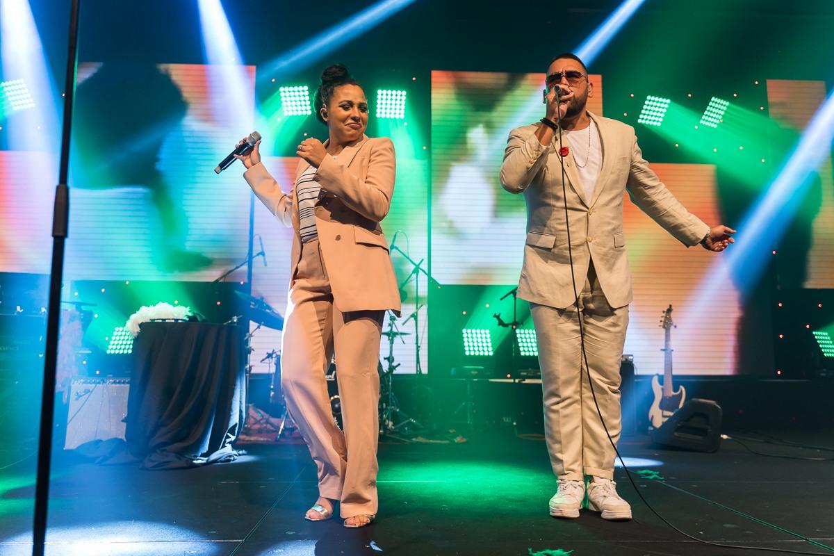 Male and female vocalists performing live on stage with microphones, colourful lighting and a full band setup behind them.