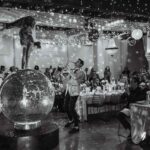 Guests watching an acrobat perform on a giant mirrored disco ball during a live floor show with saxophone and dancers.