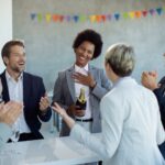 Colleagues applauding a smiling woman holding a champagne bottle during a workplace celebration.