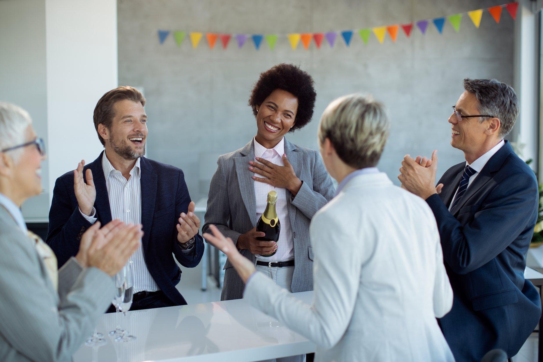 Retirement-Party-Ideas Colleagues applauding a smiling woman holding a champagne bottle during a workplace celebration.