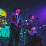Two male musicians singing and playing bass and electric guitar on stage under colourful purple and green lighting.