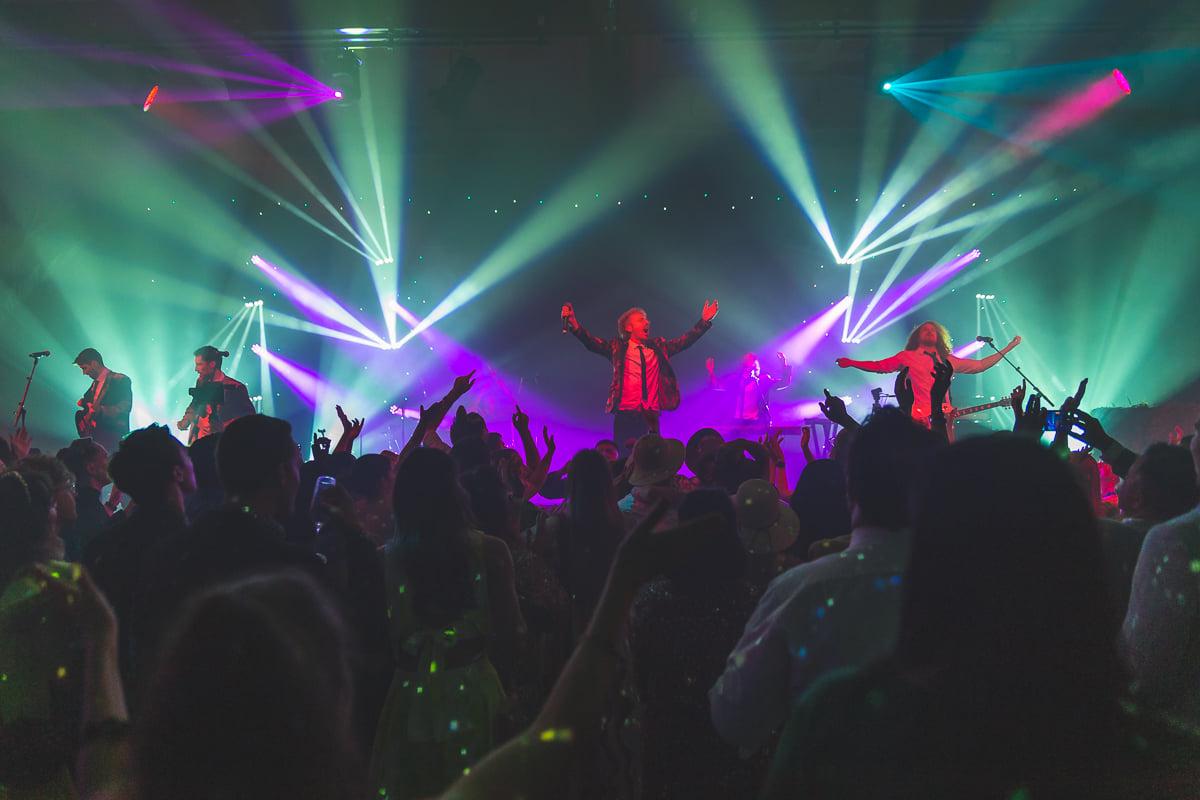Energetic live band performing under colourful concert lighting as audience members raise their hands towards the stage.